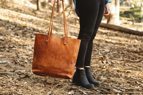 A person stands outdoors on leaf-covered ground, wearing black pants and boots, holding the Rogue Industries Fore Street Tote Bag—this stylish leather tote completes the autumn look.