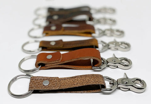 A close-up of six Rogue Industries Leather Key Chains with metal clasps, arranged in a line on a white surface in various brown shades, with the nearest keychain sharply in focus.