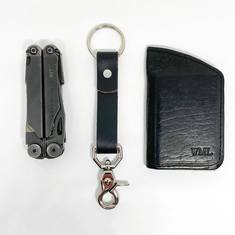 A folded Leatherman multitool, a Rogue Industries Leather Key Chain with metal clip, and a black leather wallet embossed with WML initials are arranged side by side on a white background.