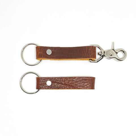 Two Rogue Industries Leather Key Chains, crafted from durable brown top-grain cowhide with silver hardware on white. The top keychain has a ring and lobster clasp; the bottom has a ring only. Both feature a metal rivet securing the loop.