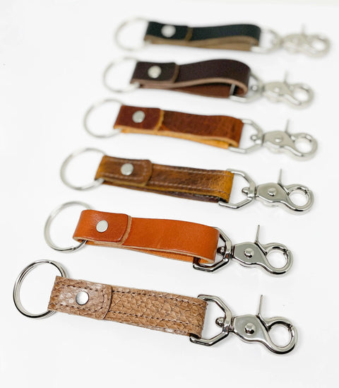 Six Rogue Industries Leather Key Chains with metal clasps and rings are displayed in a row on a white background, each crafted from durable top-grain cowhide in assorted brown or black shades and textures.