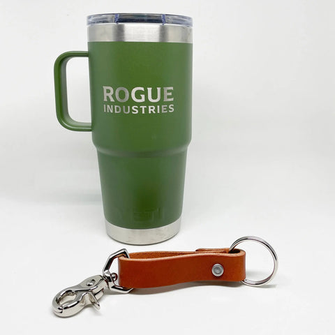 A brown Leather Key Chain from Rogue Industries, made of durable top-grain cowhide with a metal keyring and clasp, sits in front of a green travel mug bearing the Rogue Industries logo on a plain white background.