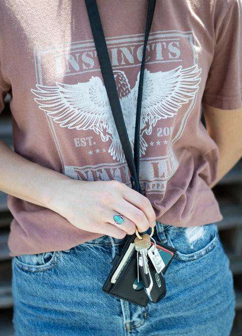 A person in a mauve eagle graphic tee and denim jeans holds keys, a wallet, and an American-made Leather Lanyard by Urban Southern. They also wear a turquoise ring.