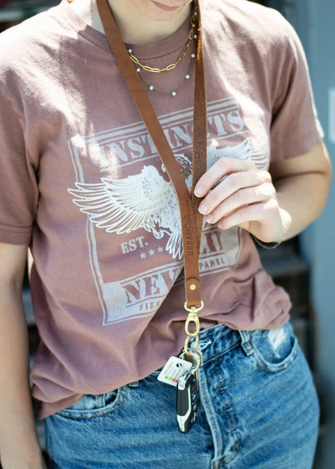 A person in a mauve graphic tee and blue jeans wears the Urban Southern Leather Lanyard with keys around their neck. The cropped image highlights the outfit and the American-made, full-grain leather accessory.