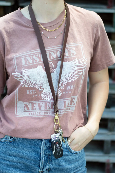 A person wearing a mauve eagle graphic t-shirt, blue jeans, and an Urban Southern Leather Lanyard holding keys and a key fob, with one hand in their pocket.