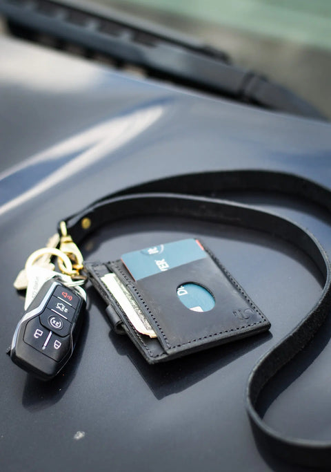 An Urban Southern Leather Lanyard displays a car key and a black full-grain leather card holder with visible cards, resting on the surface of a dark-colored vehicle.
