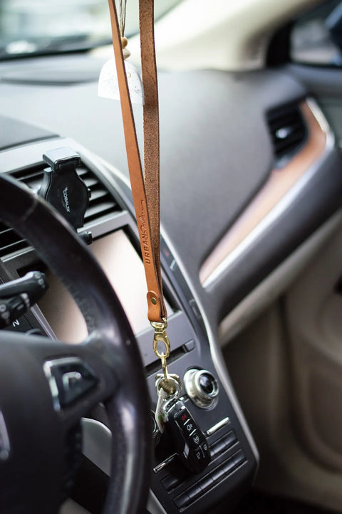 A close-up of a car’s interior shows the dashboard and steering wheel, with car keys and an Urban Southern Leather Lanyard hanging from the ignition. A phone mount is attached to the modern dashboard.