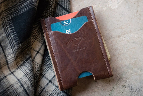 A brown leather Money Clip Wallet by Urban Southern, featuring visible stitching and holding a blue and red card, rests on a beige surface beside black and white plaid fabric. Urban Southern is embossed on the wallet.