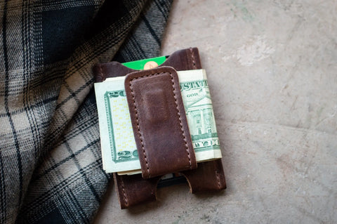 The Urban Southern Money Clip Wallet, crafted in brown leather, holds folded bills and cards and rests on a textured surface beside black and white plaid fabric.
