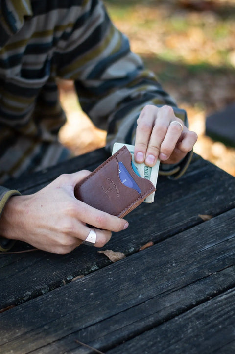 A person in a striped shirt pulls a folded hundred-dollar bill from an Urban Southern Money Clip Wallet while seated at a weathered wooden outdoor table.