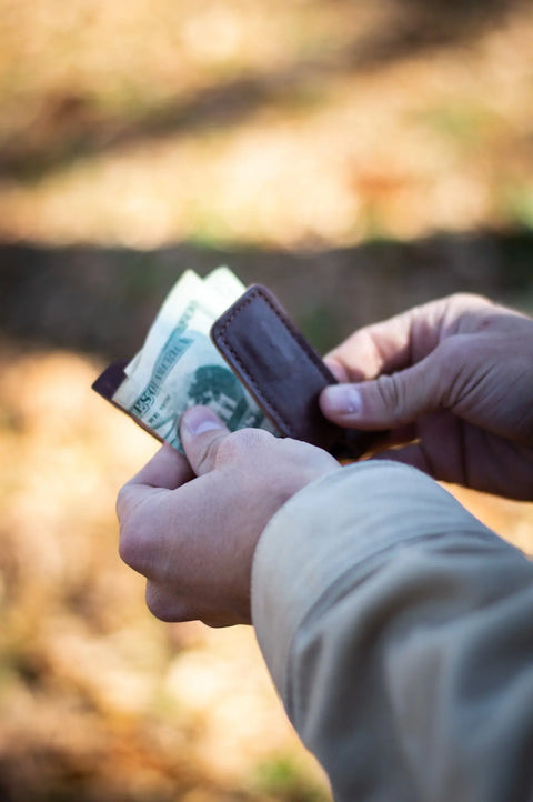 A person holds an Urban Southern Money Clip Wallet with several US dollar bills outdoors, set against a blurred natural background.