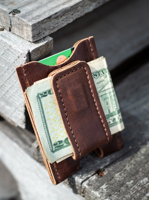The Urban Southern Money Clip Wallet, crafted in brown leather, holds a folded twenty-dollar bill and a green card, displayed on a weathered wooden surface.