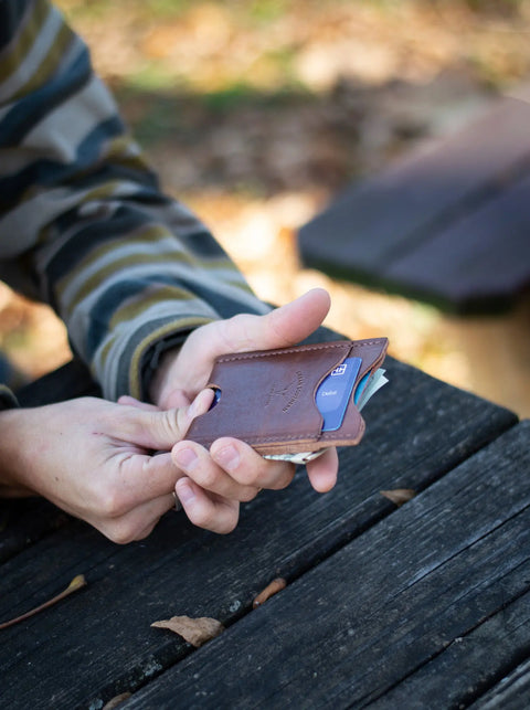 Seated at a wooden outdoor table with fallen leaves, a person in a striped long-sleeve shirt holds the Urban Southern Money Clip Wallet, slim with visible credit cards.