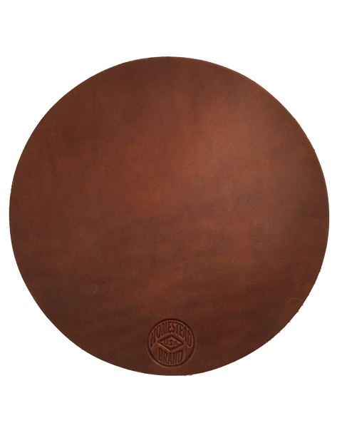 The Leather Pot Holder No. 2001 by Homestead Brand is a round, full grain leather pot holder with a smooth surface and a stamped Homestead Brand USA logo near the edge, offering stylish protection for your table.