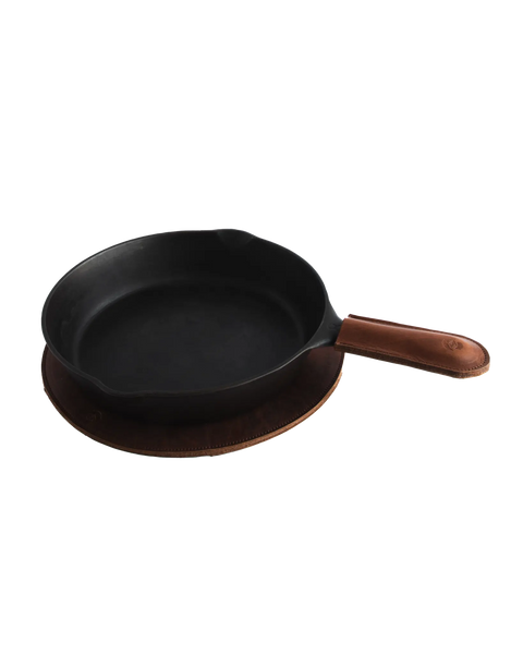 The Homestead Brand Leather Pot Holder No. 2001 sits beneath a cast iron skillet with a brown full grain leather handle cover, offering stylish heat protection on a white background.