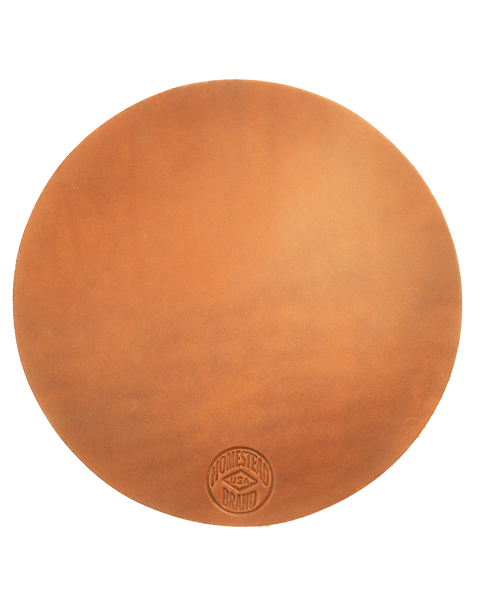 A round, light brown full grain leather pot holder—Leather Pot Holder No. 2001 by Homestead Brand—features a stamped logo near the bottom edge, shown on white. Perfect for stylish table protection.