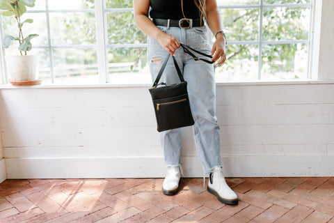 A person stands indoors by large windows and a potted plant, wearing urban slim light blue ripped jeans, a black top, white boots, a black belt, and holding the Urban Slim Crossbody by Urban Southern.