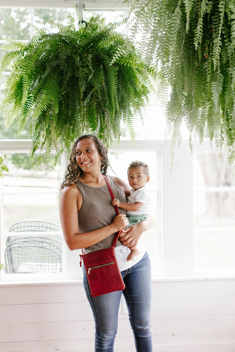 A woman in a gray sleeveless top and jeans stands indoors by large windows with ferns above, holding a young child. She carries the Urban Slim Crossbody from Urban Southern, and both look toward the camera.