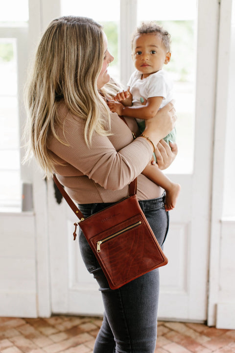 A woman with long, blonde hair holds a baby while standing indoors by a bright window. She wears a brown top, jeans, and carries the Urban Southern Urban Slim Crossbody bag. The baby looks at the camera.
