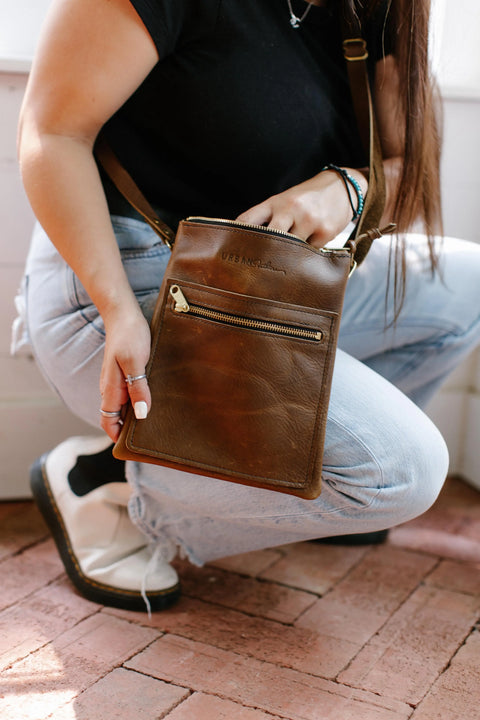 A person in light blue jeans and a black top kneels on a brick floor, holding the Urban Southern Urban Slim Crossbody bag in brown leather with a front zipper pocket.