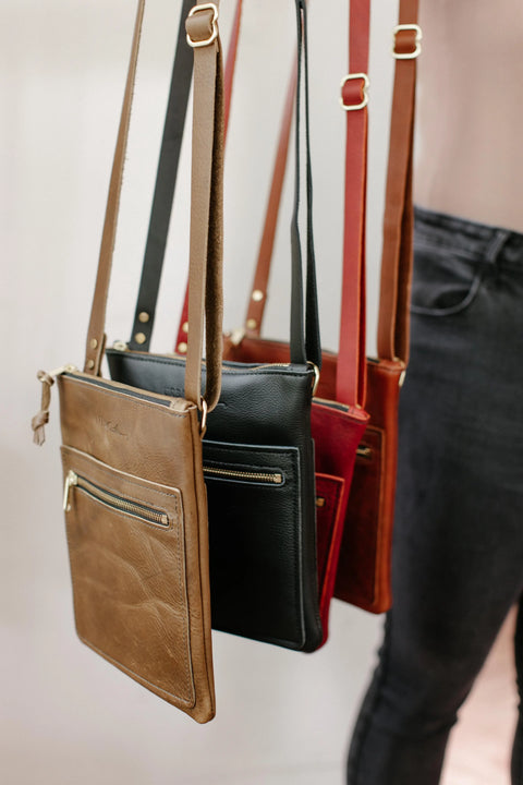 Four Urban Southern Urban Slim Crossbody bags in tan, black, red, and brown hang by their straps. Each lightweight leather bag features a front zipper pocket. A person in dark pants stands beside them against a light background.