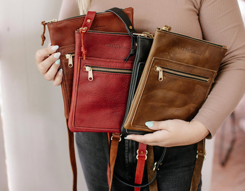 A person in a beige long-sleeve top holds several Urban Southern Urban Slim Crossbody bags in brown, red, and black, each with a front zipper pocket and gold hardware.