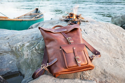 The Urban Southern Urban Rucksack rests on a large rock by the river, with a canoe nearby and a small campfire glowing in the background.