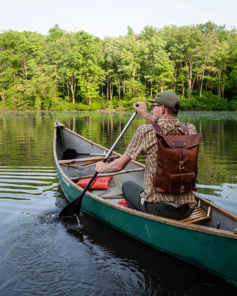 A person with an Urban Southern Urban Rucksack paddles a green canoe on a calm lake, surrounded by lush trees on a sunny day.