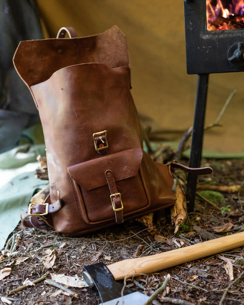The Urban Southern Urban Rucksack, crafted from full-grain leather with sturdy buckles, rests on the forest floor beside an axe and a glowing black wood stove. Twigs and leaves complete this rustic woodland scene.