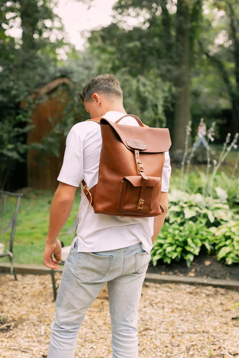 A person in a white t-shirt and light jeans stands outdoors, facing away with the Urban Southern Urban Rucksack, crafted from full-grain leather, on their back. Lush green plants and trees in the background evoke a sense of adventure.