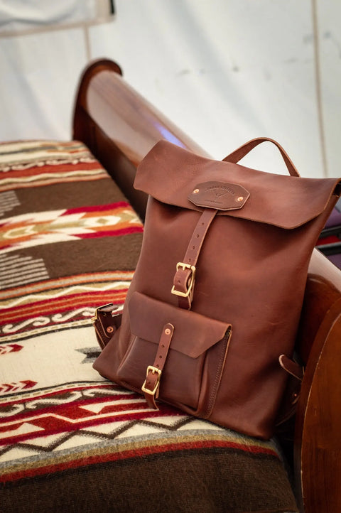 The Urban Southern Urban Rucksack, crafted from full-grain leather with gold buckles, sits atop a red, white, and brown patterned blanket on a wooden bed frame, creating a warm and inviting scene.