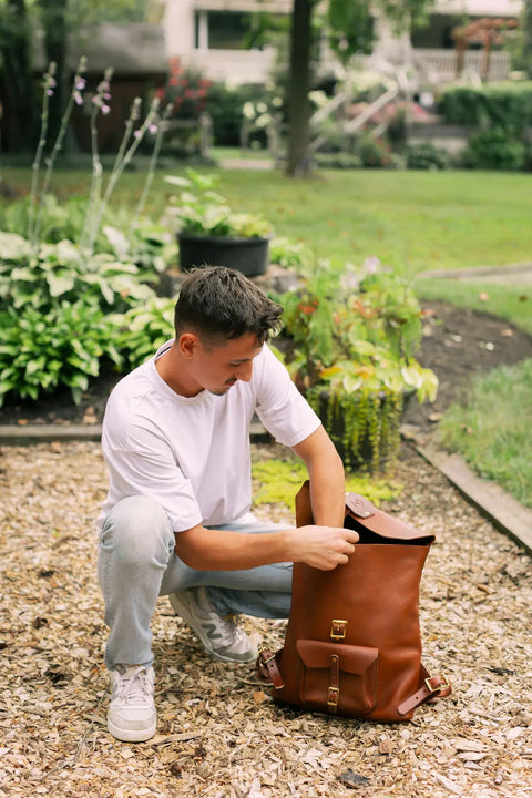 A man in a white t-shirt and light jeans kneels on a path, reaching into an Urban Southern Urban Rucksack, with green plants and a yard in the background.