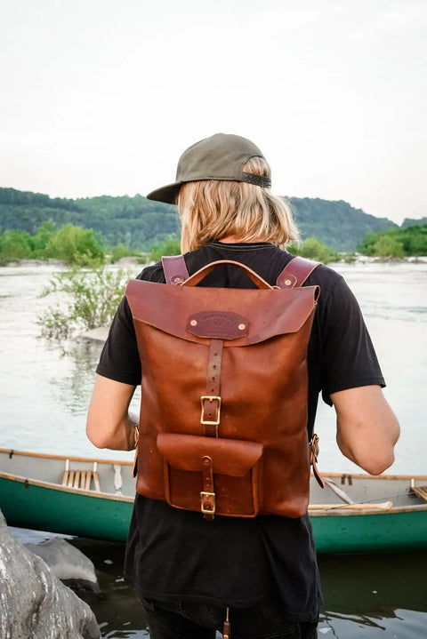 A person with long blonde hair, in a green cap and black shirt, carries an Urban Southern Urban Rucksack near a river. A green canoe and trees appear in the background as they face away.