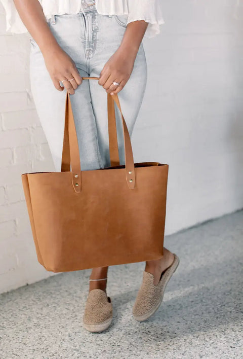 A person stands against a white brick wall, holding the Urban Southern Urban Tote by its handles and wearing light blue jeans, a white top, and beige slip-on shoes.