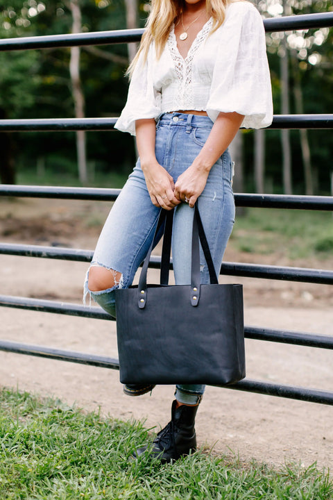 A woman stands outdoors by a black fence, holding the Urban Southern Urban Tote. She wears a white blouse, high-waisted ripped jeans, and black boots. Her face is not visible; grass and trees are in the background.