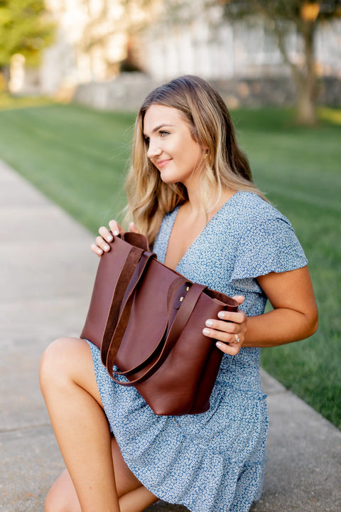 A young woman with long hair sits outdoors on a sidewalk, holding the Urban Southern Urban Tote. Shes wearing a light blue patterned dress, with green grass and blurred buildings in the background.