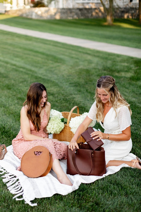 Two women enjoy a sunny picnic on a white blanket, smiling as they look into their Urban Southern Urban Tote bags. Dressed in light summer dresses, with a basket of white flowers beside them, they relax on the grass.
