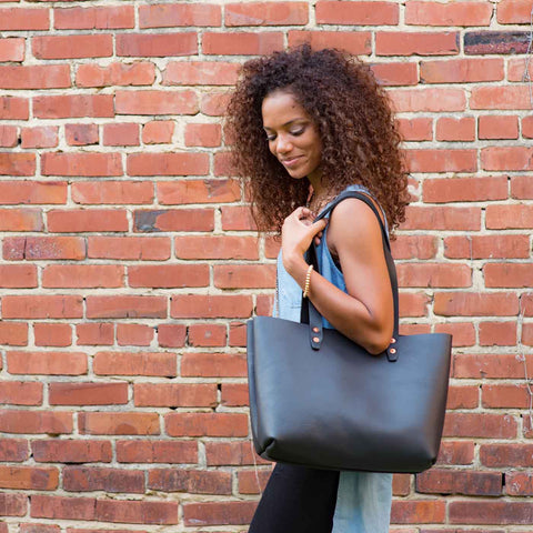 A woman with curly hair stands before a brick wall, smiling softly as she carries the Urban Southern Urban Tote over her shoulder, styled with a sleeveless denim top and black pants.