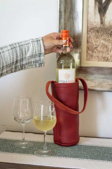 A hand places a bottle of Stella Rosa wine in an Urban Southern Urban Wine Tote, handcrafted in the USA, next to two glasses of white wine on a table, with a framed picture hanging in the background.