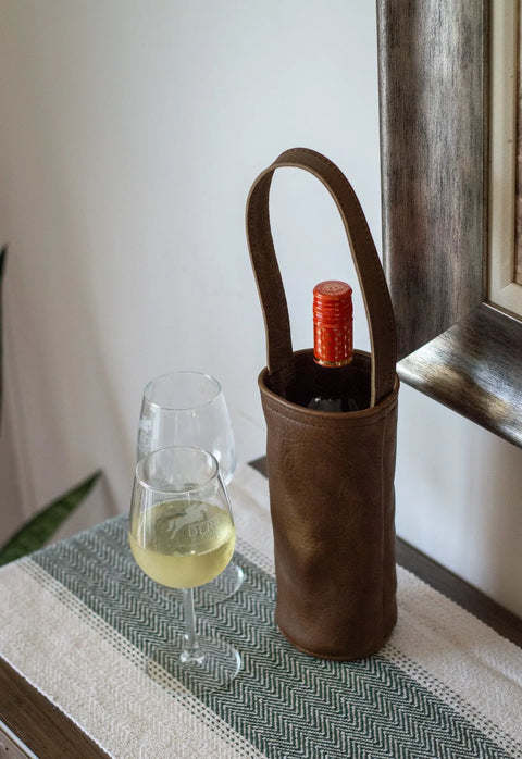 The Urban Southern Urban Wine Tote, handcrafted in brown leather in the USA, holds a bottle on a table with two filled wine glasses and a framed mirror atop a green and white patterned runner.