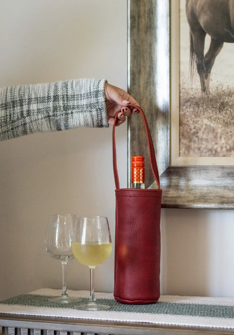 A hand holds the Urban Southern Urban Wine Tote—crafted in the USA with red leather and a wine bottle inside—beside two wine glasses (one with white wine) on a table beneath a framed picture.