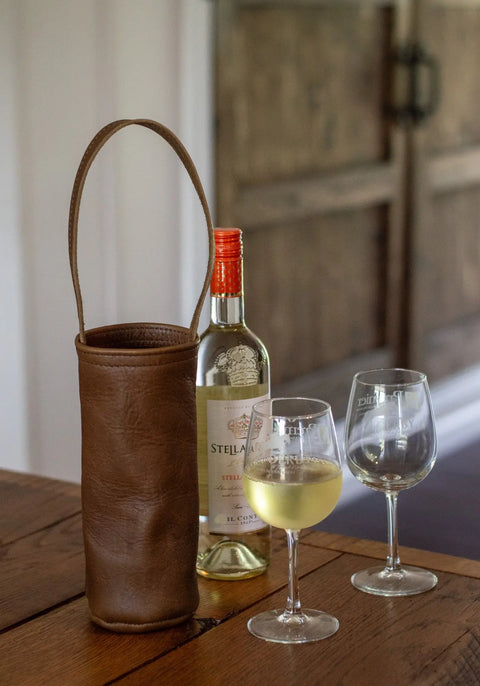 A bottle of white wine with an orange cap sits on a wooden table next to the Urban Southern Urban Wine Tote and two wine glasses, one partially filled and the other empty.