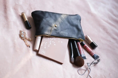 The Urban Southern Zipper Pouch, a black leather makeup bag with a gold zipper, lies open on a pink surface with cosmetics, brushes, lipsticks, an eyelash curler, and gold jewelry spilling out.