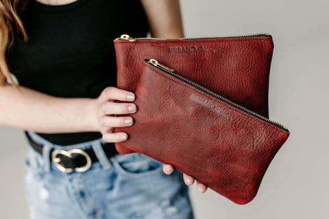 A person in a black top and denim shorts holds two Urban Southern Zipper Pouches—red leather with gold zippers—against a neutral background.