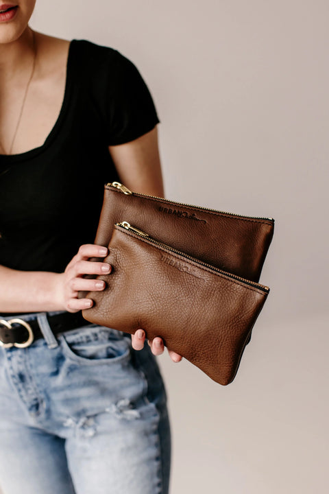 A person in a black t-shirt and ripped jeans holds two brown Urban Southern Zipper Pouches, each with a sleek gold zipper closure, against a plain background.