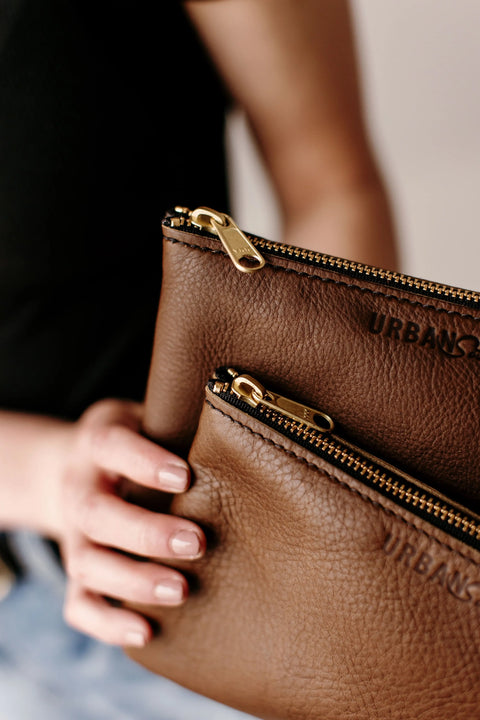 A person holds two brown leather Zipper Pouches from Urban Southern, each with a gold zipper and Urban Southern embossed; only their hand, black shirt, and blue jeans are visible.