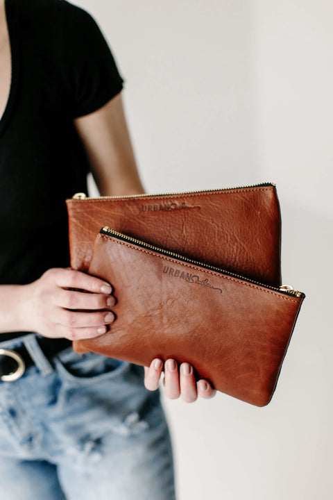A person in a black shirt and blue jeans holds two brown Urban Southern Zipper Pouches with gold zipper closures and embossed branding, set against a plain light background.
