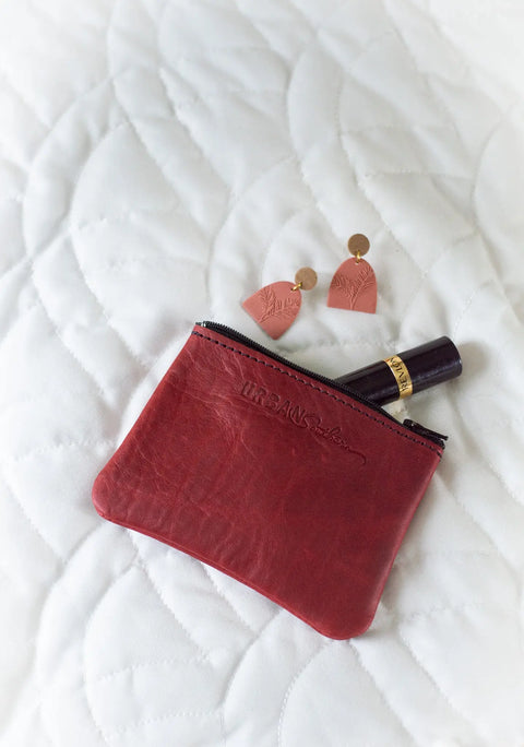 An Urban Southern Zipper Pouch, pink and gold earrings, and a dark lipstick tube are displayed on a white quilted surface.