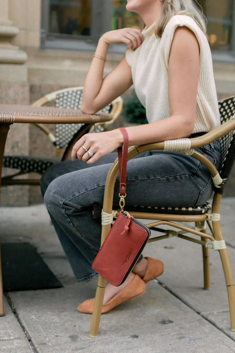 A woman at an outdoor café wears a sleeveless cream top, gray jeans, and tan flats. On her wrist hangs the Urban Southern Urban Zipper Wallet, a red leather wallet with an accordion-style design and credit card slots.