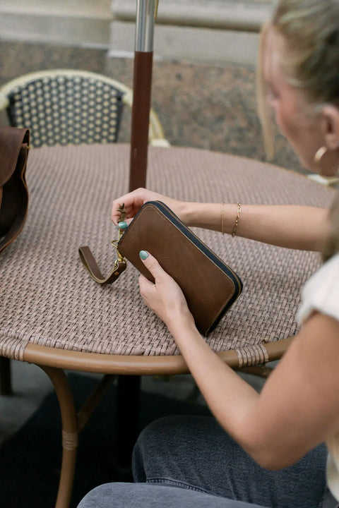 A person with light nail polish holds the Urban Southern Urban Zipper Wallet at an outdoor woven table next to a matching brown bag, creating a relaxed and casual vibe.
